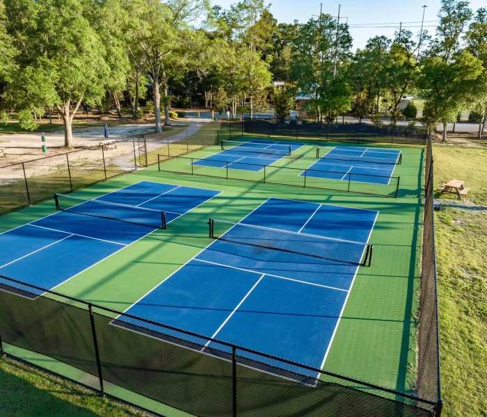 An aerial view shows a fenced-in pickleball complex at Splash RV Resort with six blue courts bordered by green surfacing, surrounded by lush trees, picnic tables, and grassy areas under bright, sunny skies.