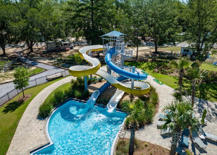 An aerial view captures a vibrant water park area at Splash RV Resort, featuring twisting yellow and blue waterslides spiraling down into a curved turquoise splash pool, surrounded by paver walkways, palm trees, and lounge chairs. In the background, RVs are parked among tall pine and oak trees under bright, sunny skies, creating a lively yet relaxing resort atmosphere.