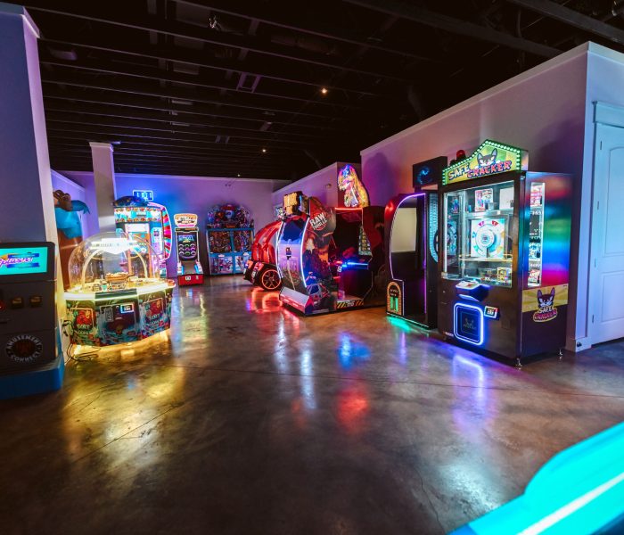 Indoor arcade with brightly lit game machines including Safe Cracker prize machine and ride-on game on glossy polished concrete floor reflecting neon pink blue and yellow glows under moody colorful lighting at Splash RV Resort in Milton, Florida.