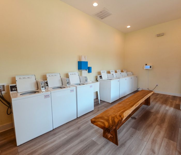 Communal laundry room with pale yellow walls light wood-look vinyl flooring white top-loading washing machines and stacked washer dryer with live-edge wooden bench illuminated by warm recessed ceiling lights at Splash RV Resort in Milton, Florida.