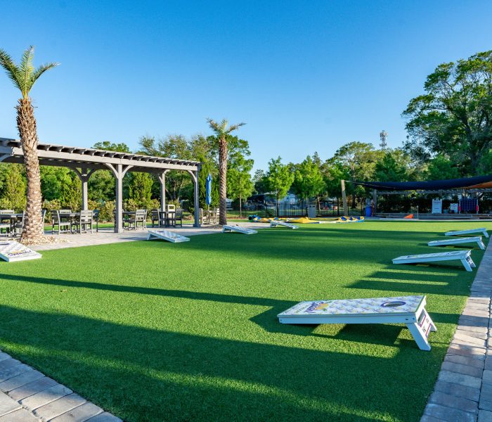 A spacious outdoor recreation area at Splash RV Resort featuring bright green artificial turf lined with white cornhole boards, bordered by paver walkways and a gray wooden pergola shading patio seating beside two palm trees.