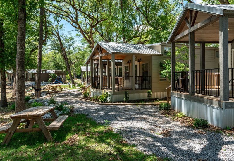 Beautifully appointed luxury cottage with soft gray exterior, vaulted covered porch, and wooden beams set among towering shade trees at Splash RV Resort in Milton, Florida.