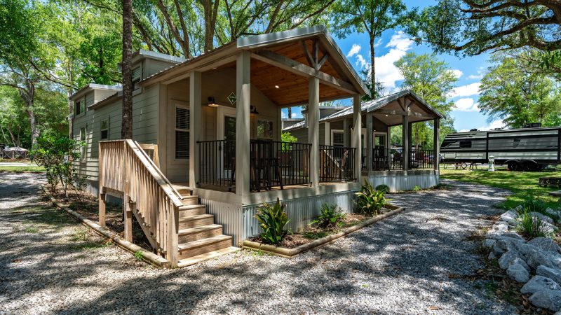 Charming sage-green luxury cottage with vaulted wood-beamed covered porch on a gravel pad surrounded by lush oak trees at Splash RV Resort in Milton, Florida.