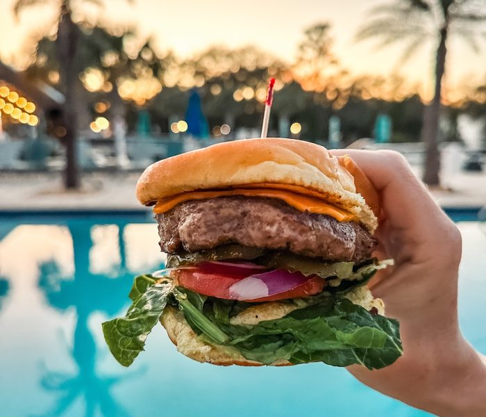 Gourmet burger held up against sparkling turquoise pool at Splash RV Resort in Milton, Florida during sunset