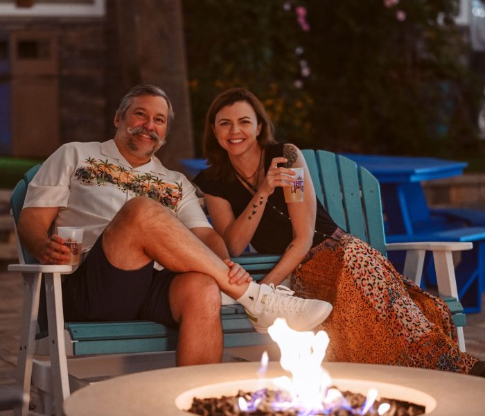 A smiling couple relaxes together on a turquoise Adirondack chair beside a modern concrete gas fire pit with dancing blue and orange flames at Splash RV Resort in Milton, Florida, with white and gold balloons and warm evening lighting creating a festive, intimate atmosphere.