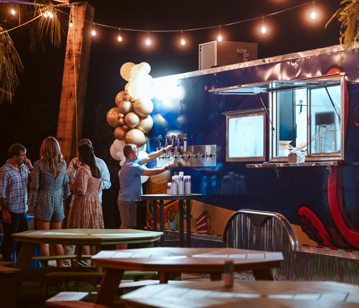 A nighttime gathering at Splash RV Resort in Milton, Florida features guests mingling around a vibrant blue food and beverage truck adorned with red octopus tentacle artwork and a row of beer taps, where a man in a blue polo pours a drink beneath glowing string lights and a cluster of gold and white balloons.