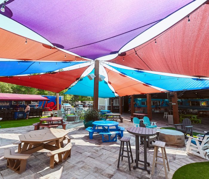 This vibrant outdoor gathering area at Splash RV Resort in Milton, Florida features a stunning canopy of overlapping purple, red, orange, and blue shade sails strung with cafe lights above a paver patio filled with octagonal wooden picnic tables, bright blue benches, and white Adirondack chairs.
